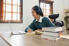 微笑女孩学生穿无线耳机研究在线老师快乐年轻的女人学习语言听讲座看网络研讨会写笔记移动PC遥远的教育