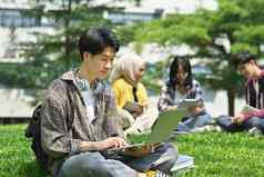 年轻的亚洲男人。学生移动PC绿色草前面大学建筑教育技术生活方式概念