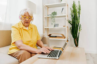 快乐上了年纪的女人移动PC打字耳机坐着首页沙发上黄色的衬衫明亮的现代室内生活方式在线沟通