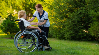 护士持有上了年纪的高加索人女人轮椅手支持护士走病人公园