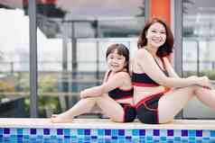 微笑亚洲妈妈。女儿坐着边缘游泳池泳衣相机