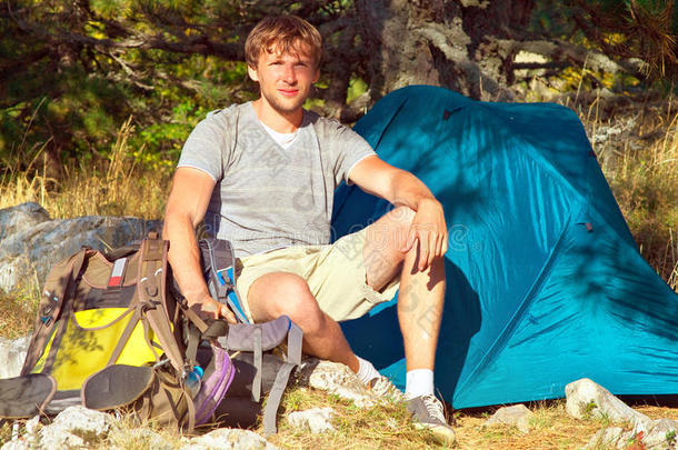 年轻男子笑脸徒步旅行者坐在背包和帐篷<strong>户外露营</strong>