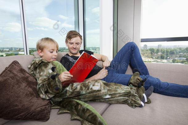 穿着恐龙服装的男孩和父亲在家沙发床上看图画书