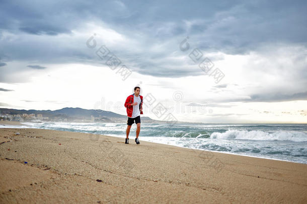 穿着红色<strong>风衣</strong>的运动型年轻跑步者在美丽的大海背景下沿着海滩奔跑