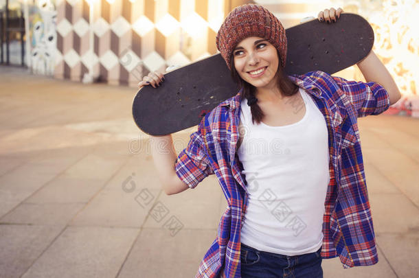 黑发少女穿着时髦的服装（牛仔裤短裤、短裤、格子衬衫、帽子）在公园户外玩滑板