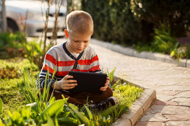 可爱的男孩坐在公园的草地上，在耳朵里玩平板电脑