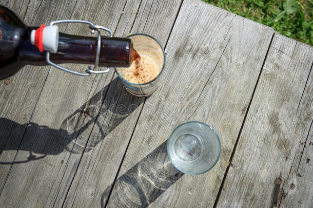 泡沫的深色啤酒从夏天花园里的<strong>棕色玻璃瓶</strong>中倒入高大的玻璃杯中，放在乡村的木桌上