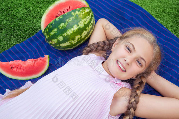 夏天可爱的小女孩在草地上吃西瓜。 马尾辫，长发，牙齿微笑，坐在草地上，享受着乐趣