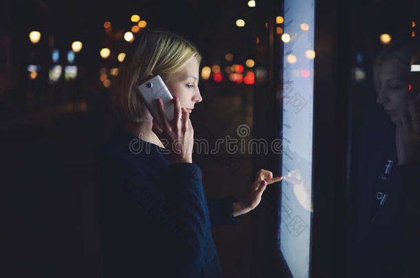 美丽的金发女郎在触摸反射光线的大数字屏幕时用手机说话，