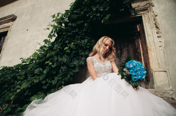美丽的年轻金发新娘，新娘的新娘花束坐在楼梯下华丽的植物