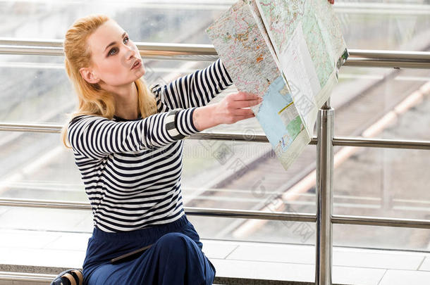 坐在车站或终点站看地图的一位穿着条纹衬衫的金发游客。