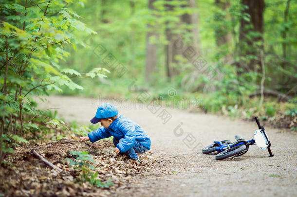 高加索小男孩穿着蓝色夹克，牛仔裤和棒球帽，在公园操场外面骑自行车，坐在地上