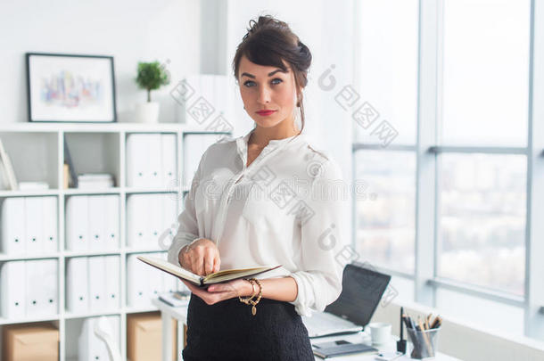 漂亮的女商人站在办公室里，拿着笔记本，计划工作日的会议，看着相机。