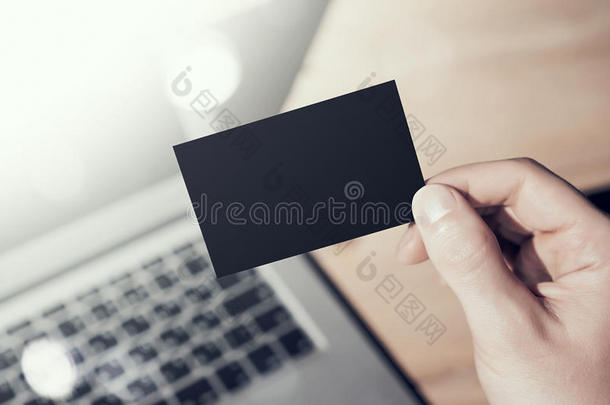 特写照片男子显示空白黑色名片，并使用现代笔记本电脑在木桌上模糊的背景。 模拟