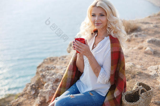 坐在岩石海岸上喝咖啡的漂亮女人
