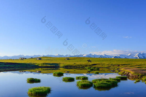 草原上有<strong>雪山</strong>，木屋，骑马人，绿色的草和它们在湖泊中的倒影。