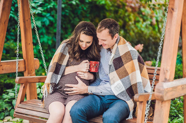 一个怀孕的年轻女人和她的丈夫。 一个幸福的家庭坐在<strong>秋</strong>千上，抱着肚子。 孕妇在公园放松。