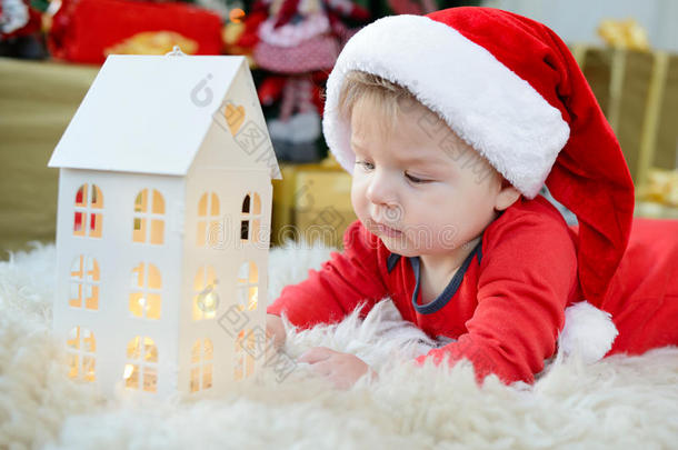 可爱的小婴儿肖像庆祝圣诞节。 新年`假期。 穿着圣诞老人服装的男孩在下面看玩具屋