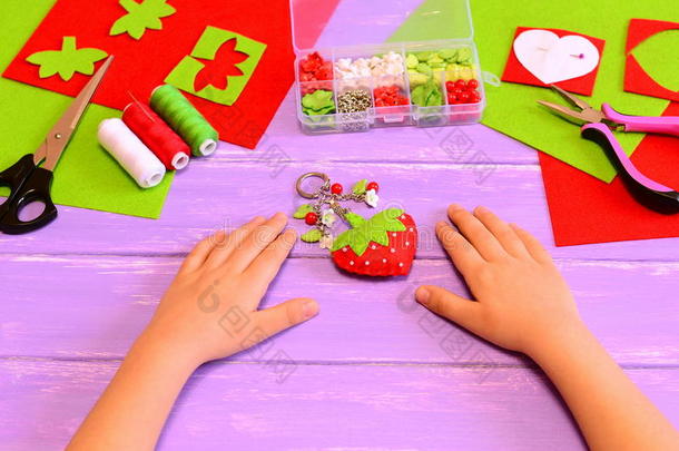孩子用毛毡和珠子做了一个草莓钥匙链。孩子展示钥匙扣工艺品。孩子们把手放在一张木桌上。手工艺品