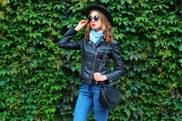 时尚的年轻女人穿着一件黑色的岩石夹克，手提包摆在绿叶墙上