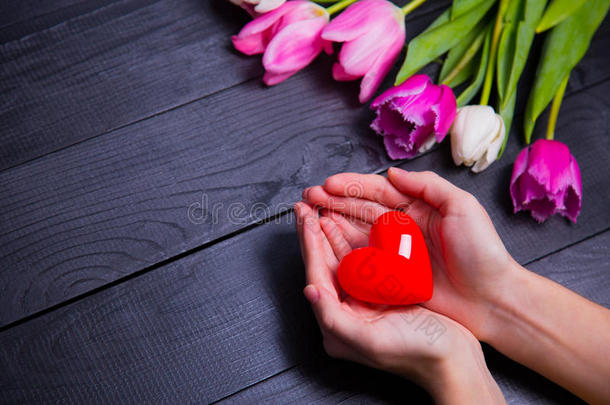 一束嫩粉色的郁金香和双手捧着红色的心在黑色的木制背景上
