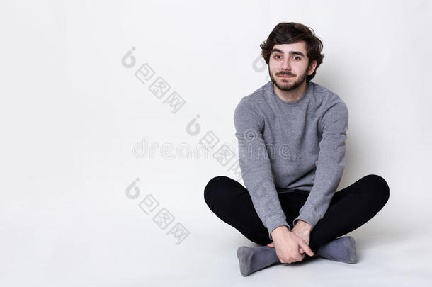 一个有着深色胡子和胡子的男人，有着时髦的发型，穿着<strong>休闲裤</strong>和灰色毛衣，坐在地板上放松