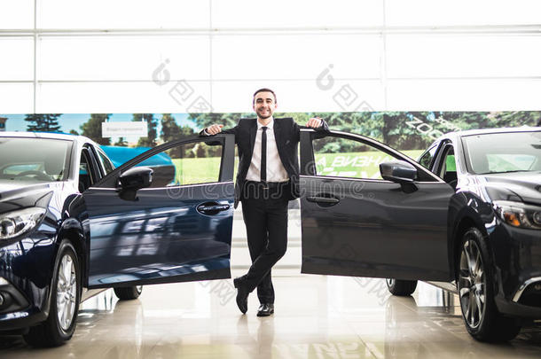 自信微笑的汽车推销员在展厅附近的两辆展厅里，他站在豪华汽车的敞开车门附近