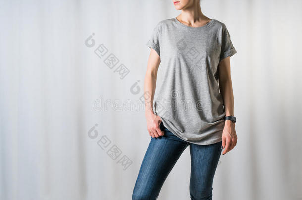 站在浅灰色背景上的年轻女子，穿着灰色的t恤和蓝色牛仔裤