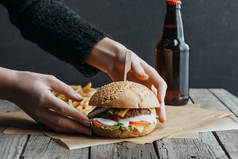 用汉堡包、法式薯条和啤酒瓶在木桌上烤纸上的女性手的裁剪视图