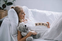 微笑的孕妇在白色睡衣与泰迪熊和木块与婴儿刻字在沙发上休息的侧面视图