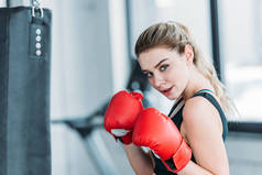 有吸引力的年轻女运动员在拳击手套看着相机在健身房