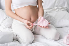 孕妇坐在床上拿着粉色婴儿衣服的裁剪视图