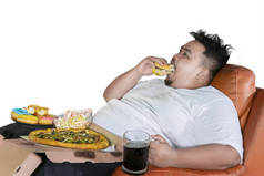 不健康的生活方式概念: 贪婪的胖子吃汉堡和垃圾食品在沙发上, 查出在白色背景