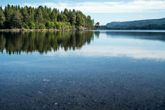 一个安静的宁静的夏日早晨, 可以看到晶莹平静的湖面上的鹅卵石, 底部的绿色森林在水面上, 地平线上有山--在大自然中的无声冥想和娱乐的概念.