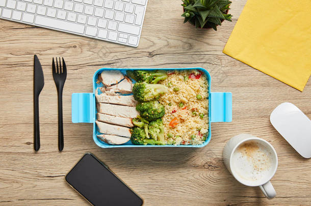午餐盒的顶视图与<strong>鸡</strong>肉，花椰菜和利索托在桌子上与电脑键盘和智能手机