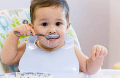 微笑的孩子在厨房吃的食物。1 年岁宝宝吃他们自己的汤。第一次的固体食物，如何教孩子用勺子吃饭。可爱的宝宝，吃午饭用勺子吃饭