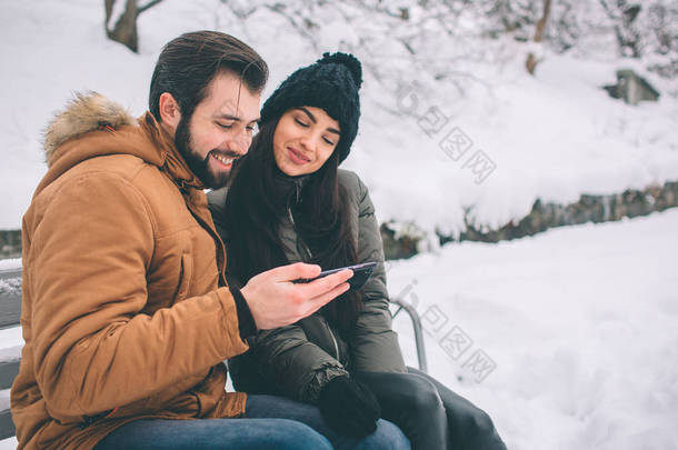 快乐的年轻夫妇在冬天。家庭户外。男人和女人看向上和笑。爱, 乐趣, <strong>季</strong>节和人-漫步在冬天公园。坐在板凳上。持有智能<strong>手机</strong>.