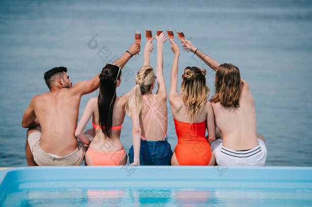 年轻男性和<strong>女性朋友</strong>的后视图坐在海滩附近池边, 无比的香槟杯