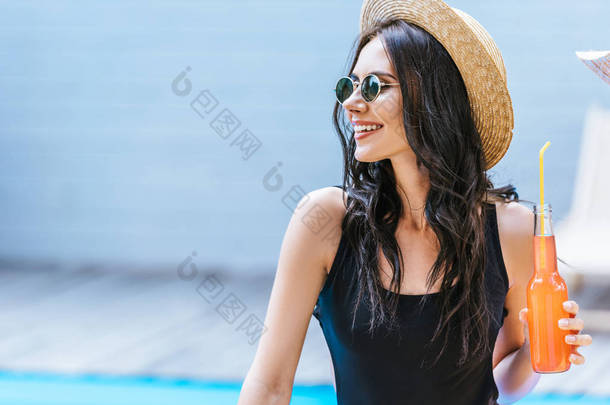 微笑的年轻妇女在泳装举行瓶与清爽的饮料和看在池畔