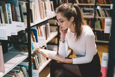 沉思的女学生在眼镜视觉保护坐在书架附近阅读有趣的小说, 聪明的集中年轻女子花时间在图书馆喜欢文学和教育