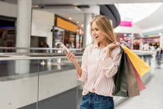 年轻微笑的女性购物者与纸袋使用智能手机在购物中心