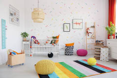 坐垫凳在斯堪婴儿卧室内的五颜六色的地毯上, 配有台灯、摇篮和海报。真实照片
