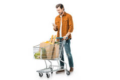 幸福英俊的人携带购物推车与产品在纸袋和使用智能手机查出的白色