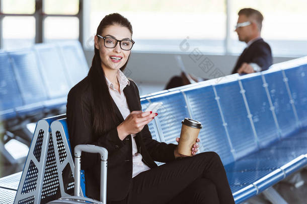 年轻的微笑的女商人在眼镜与咖啡去使用智能手机在机场候机室
