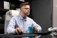 戴眼镜的商务人员在电脑桌旁的办公室工作, 喝着小杯的咖啡