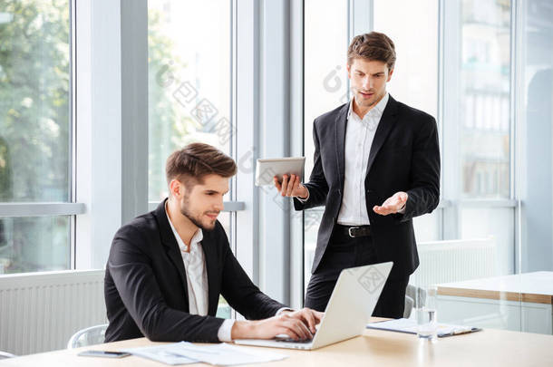 两个英俊的年轻商人在办公室使用笔记本电脑和平板电脑