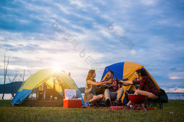 组的男人和女人享受露营野餐和烧烤在帐篷中背景的湖。年轻混合种族亚洲女人和男人。年轻人们手里烘培和欢呼瓶啤酒.
