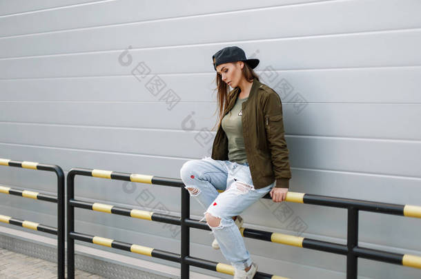 戴着棒球帽，在街头风格的衣服，坐在一堵墙附近年轻时尚美女