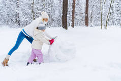 快乐的母亲和女儿在冬天公园堆雪人的侧面图