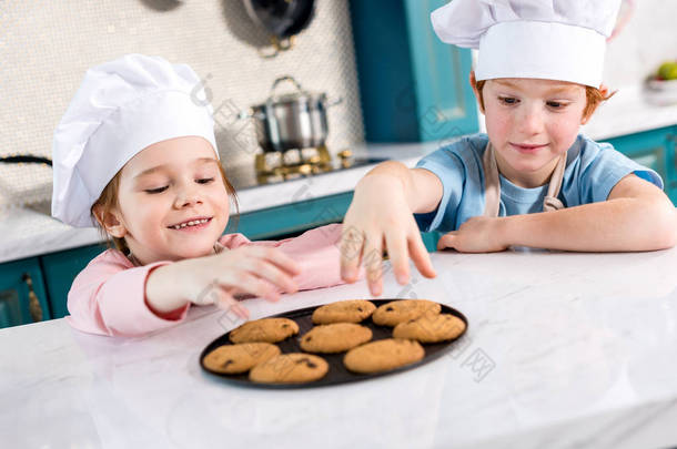 愉快的小孩子在厨师帽子吃可口曲奇饼在厨房